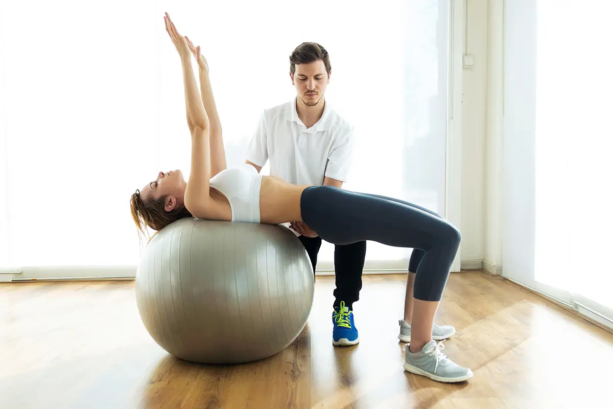 Shot of physiotherapist helping patient to do exercise on fitness ball in physio room.
